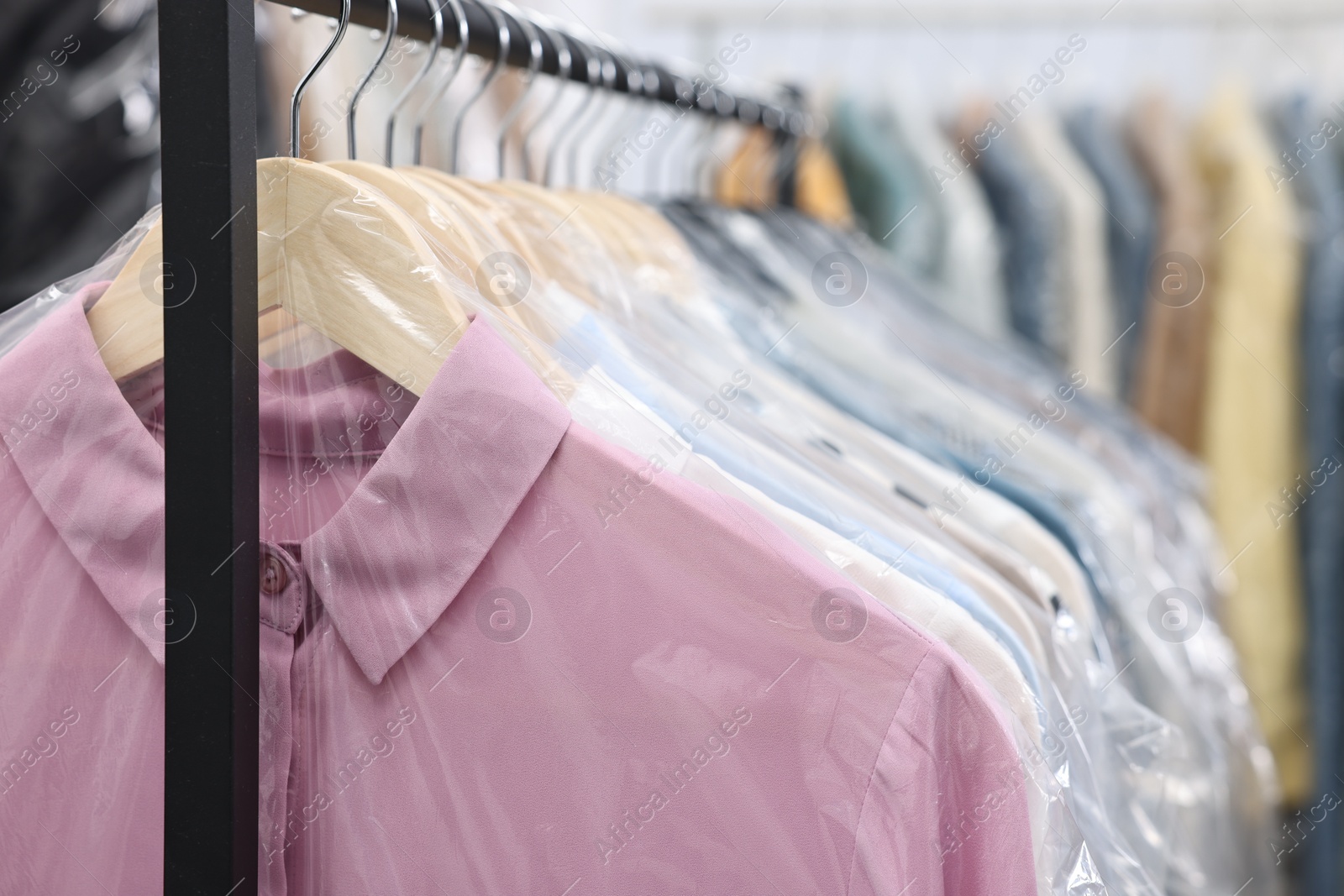 Photo of Dry-cleaning service. Many different clothes in plastic bags hanging on rack indoors, closeup