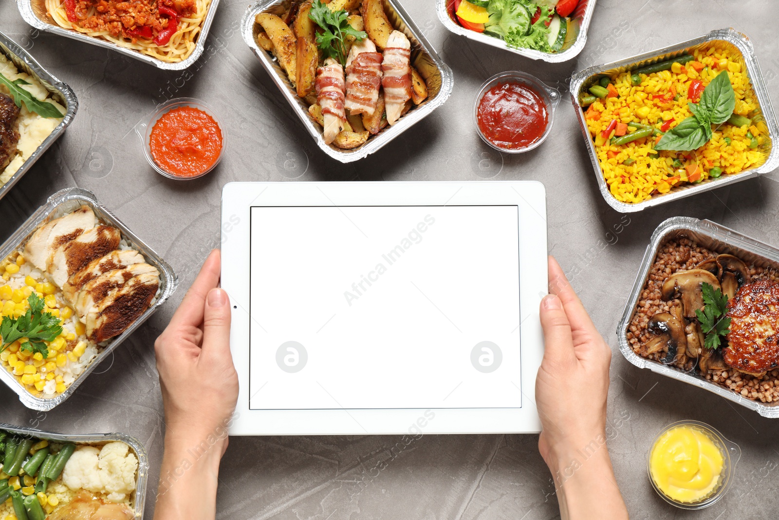 Photo of Top view of woman holding tablet over grey table with lunchboxes, mockup for design. Healthy food delivery