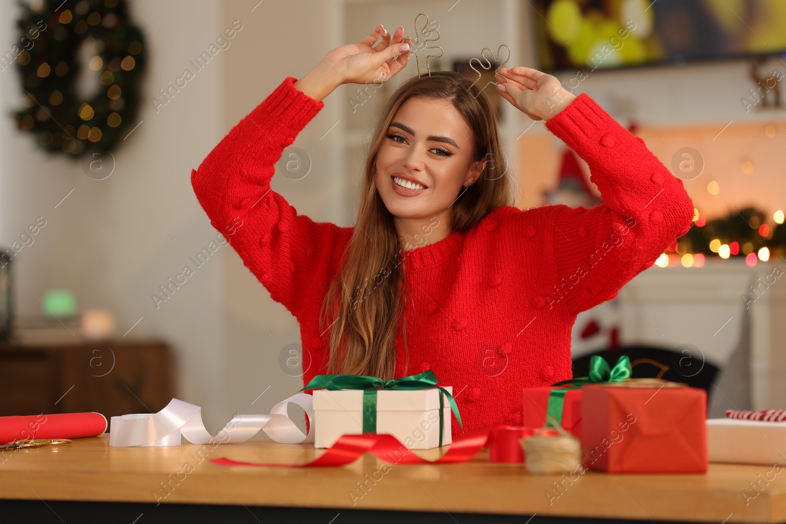 Photo of Beautiful young woman in deer headband, Christmas gifts and decor at table in room