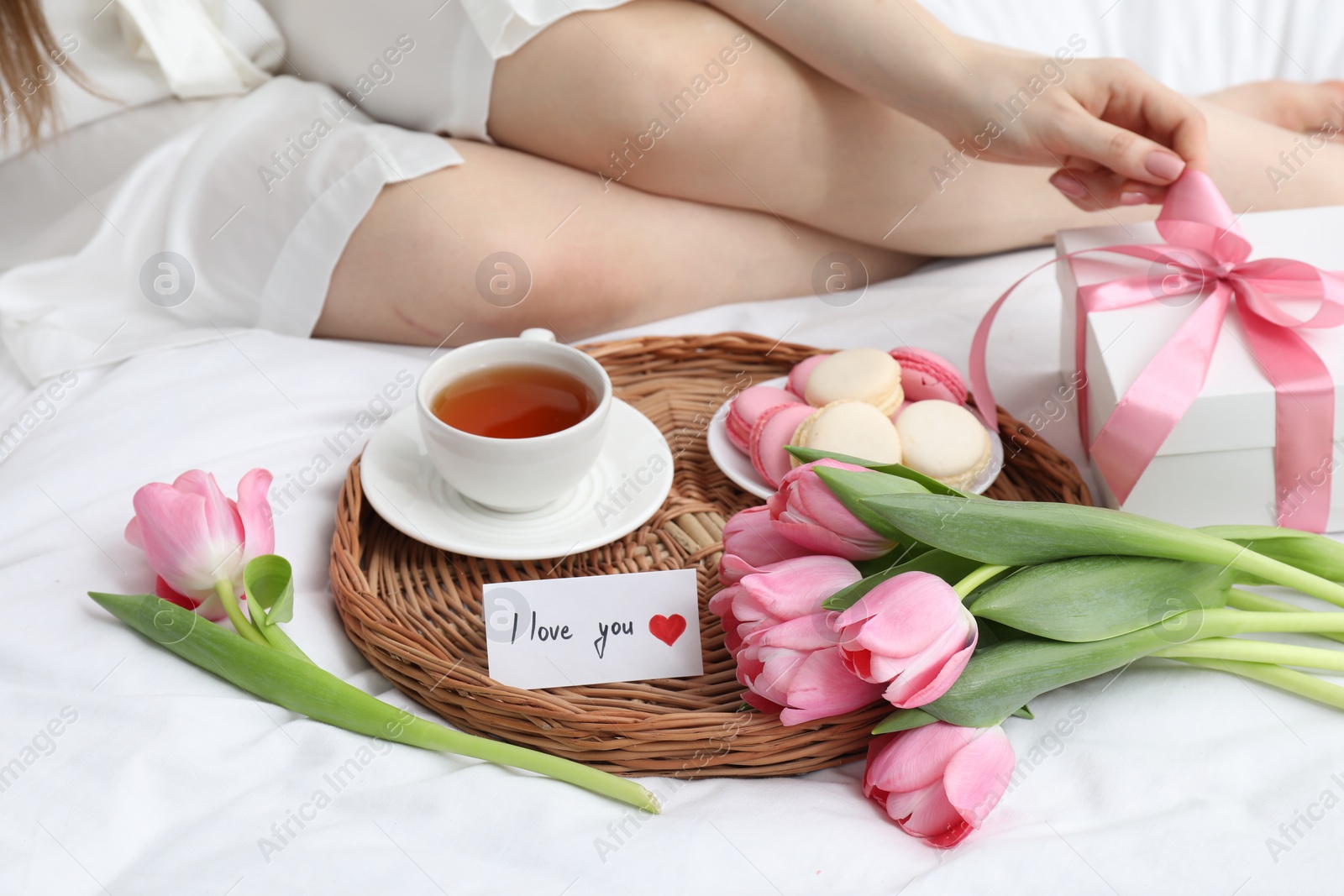 Photo of Tasty breakfast served in bed. Woman with gift box, tea, macarons, flowers and I Love You card at home, closeup