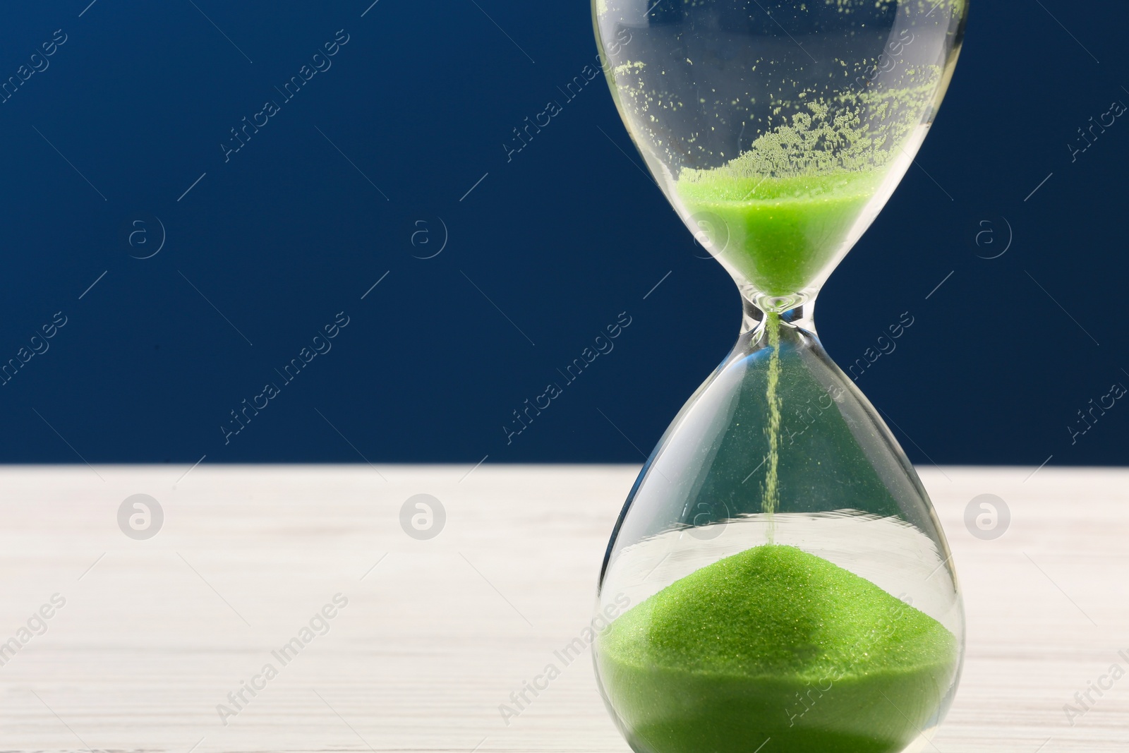 Photo of Hourglass with light green flowing sand on white table against dark blue background, closeup. Space for text