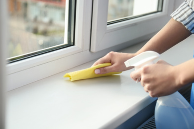 Photo of Woman cleaning window sill with rag and detergent indoors, closeup
