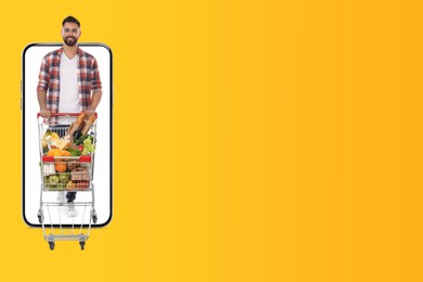 Image of Grocery shopping via internet. Happy man with shopping cart full of products walking out of huge smartphone on orange background, space for text