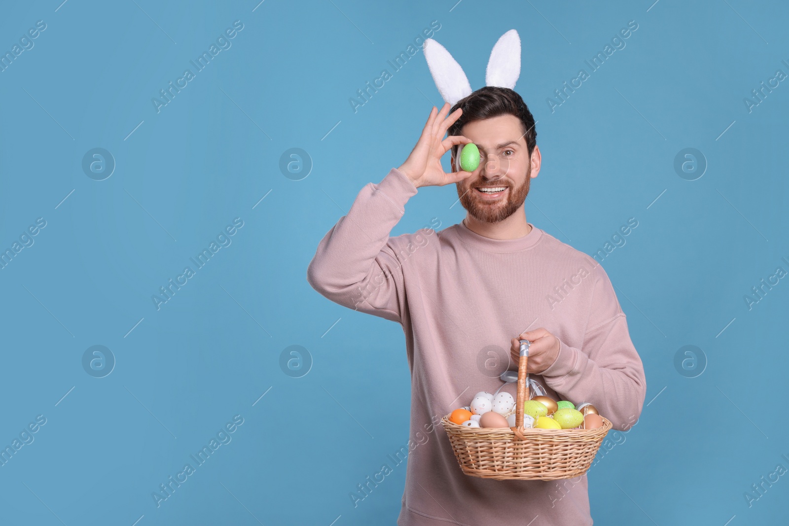 Photo of Happy man in cute bunny ears headband covering eye with Easter egg and holding wicker basket on light blue background, space for text