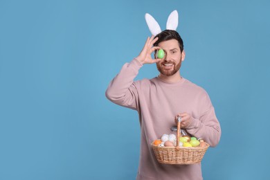 Happy man in cute bunny ears headband covering eye with Easter egg and holding wicker basket on light blue background, space for text