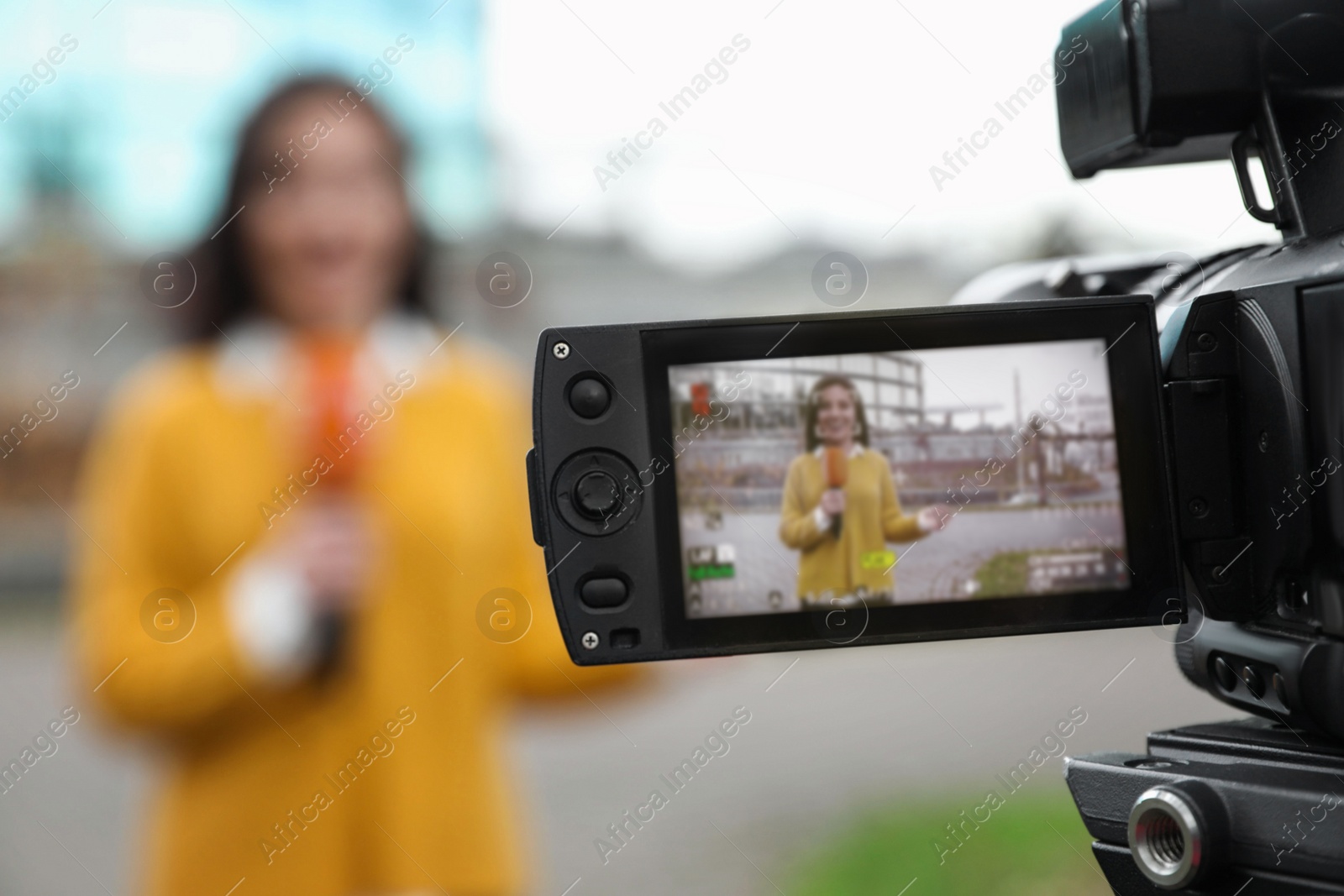 Photo of Young female journalist with microphone working on city street, focus on camera display