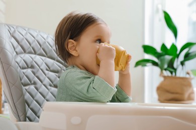 Cute little baby drinking from cup in high chair indoors