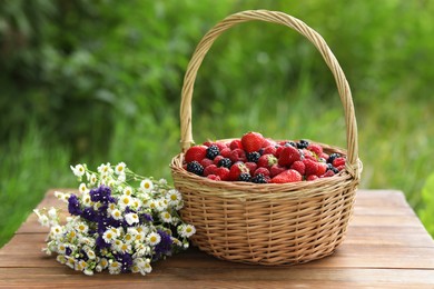 Photo of Wicker basket with different fresh ripe berries and beautiful flowers on wooden table outdoors