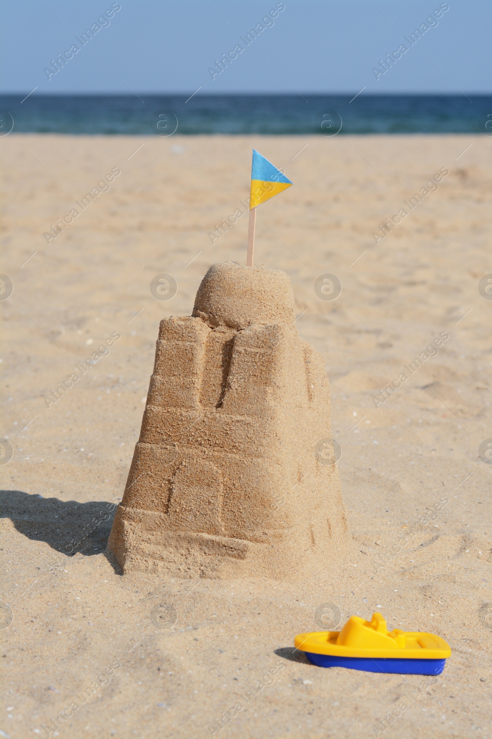 Photo of Beautiful sand castle with small Ukrainian flag and toy boat on beach near sea