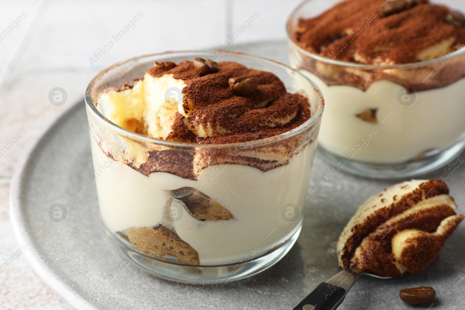 Photo of Delicious tiramisu in glasses, spoon and coffee beans on table, closeup