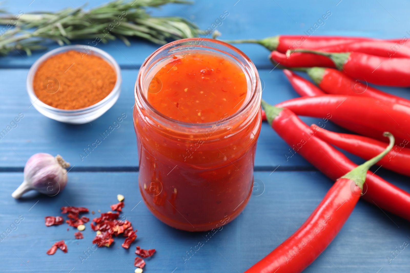 Photo of Spicy chili sauce in jar and ingredients on blue wooden table, closeup