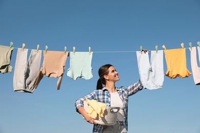 Smiling woman hanging baby clothes with clothespins on washing line for drying against blue sky outdoors