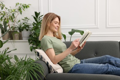 Woman reading book on sofa surrounded by beautiful potted houseplants at home