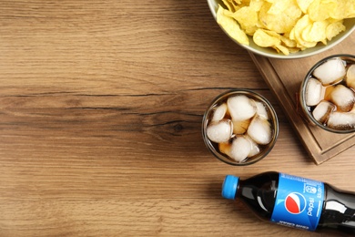 Photo of MYKOLAIV, UKRAINE - FEBRUARY 15, 2021: Glasses and bottle of Pepsi with snack on wooden table, flat lay. Space for text