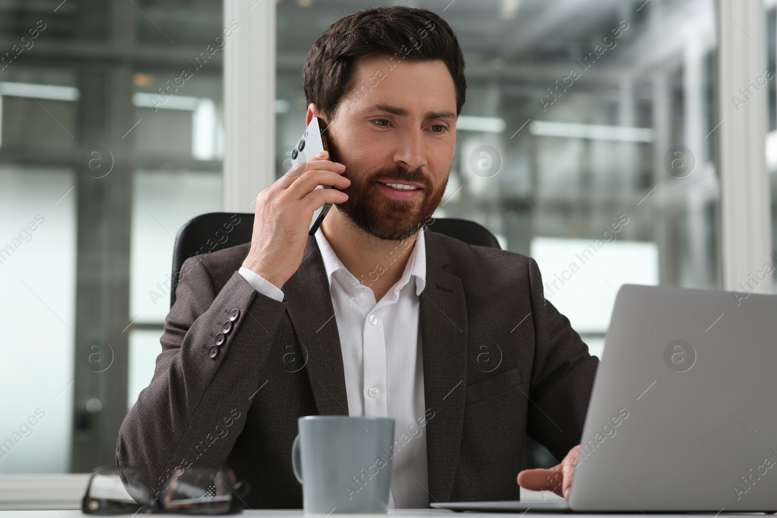 Photo of Man talking on phone while working with laptop at white desk in office