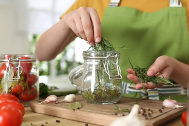 Photo of Woman putting herbs into pickling jar at table in kitchen, closeup