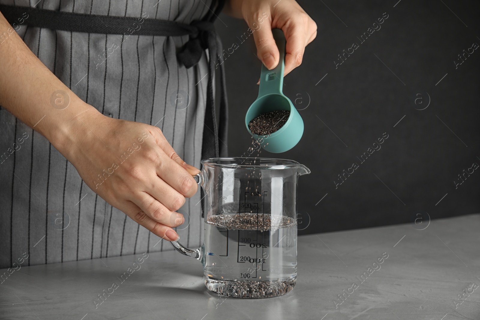 Photo of Young woman pouring chia seeds into measuring glass with water at table, closeup