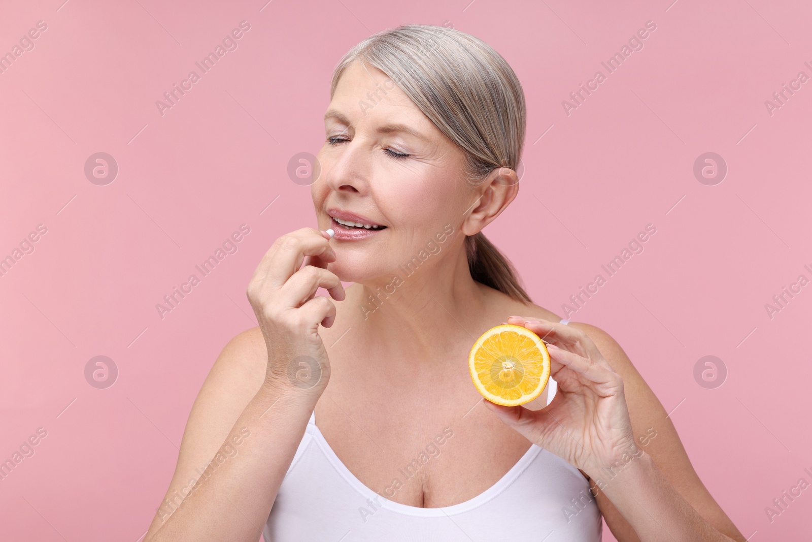 Photo of Beautiful woman with half of orange taking vitamin capsule on pink background