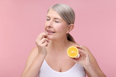 Beautiful woman with half of orange taking vitamin capsule on pink background