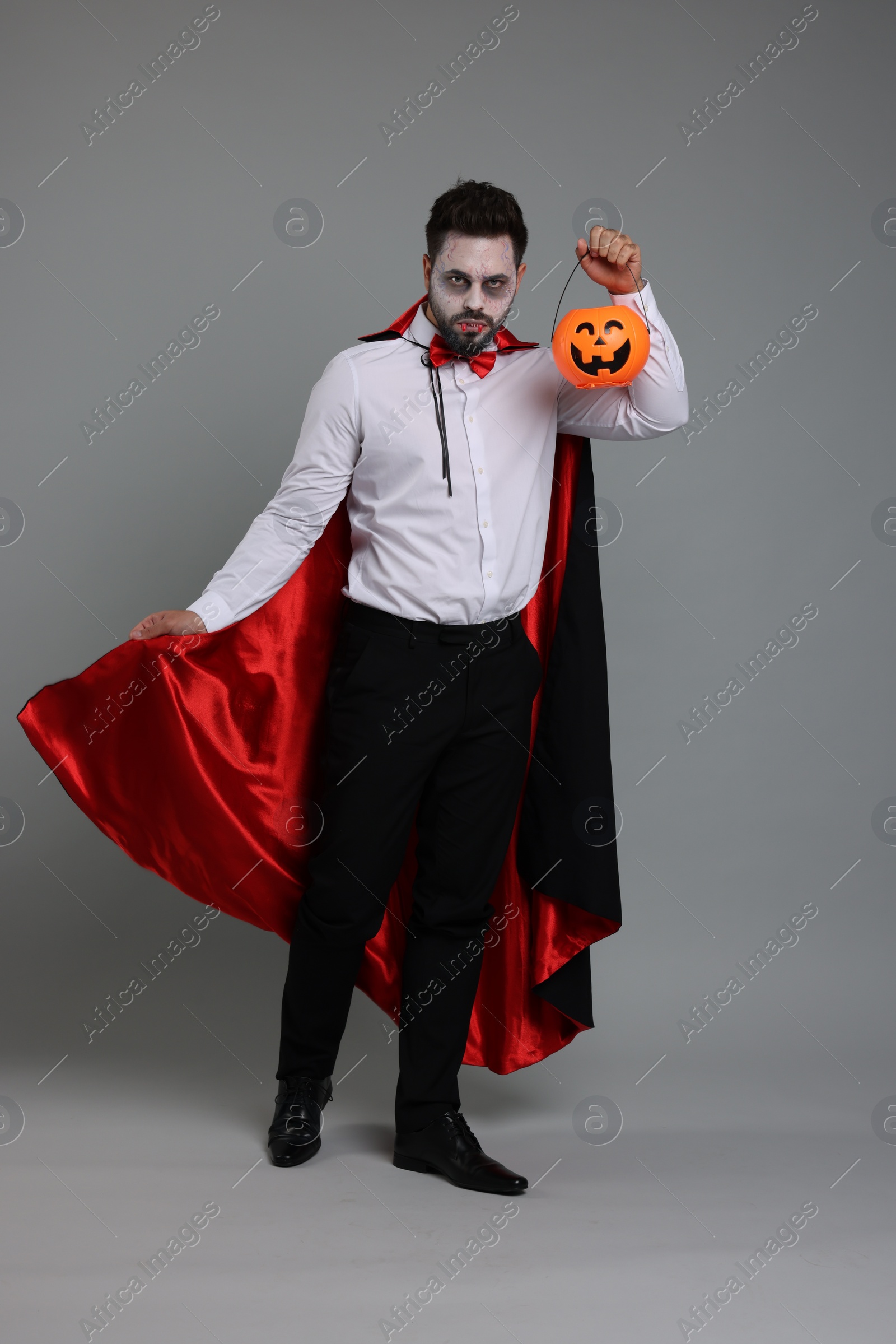 Photo of Man in scary vampire costume with fangs and pumpkin bucket on light grey background. Halloween celebration