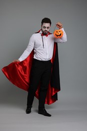 Photo of Man in scary vampire costume with fangs and pumpkin bucket on light grey background. Halloween celebration
