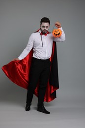 Man in scary vampire costume with fangs and pumpkin bucket on light grey background. Halloween celebration