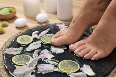 Photo of Woman soaking her feet in plate with water, flower petals and lime slices, closeup. Pedicure procedure