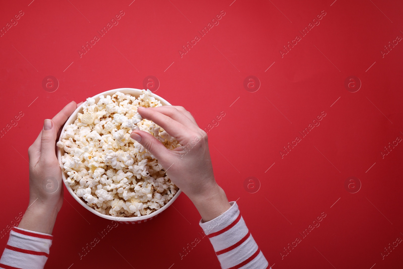 Photo of Woman taking delicious popcorn from paper bucket on red background, top view. Space for text