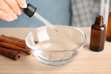 Photo of Woman dripping cinnamon essential oil from pipette into bowl of water on table, closeup