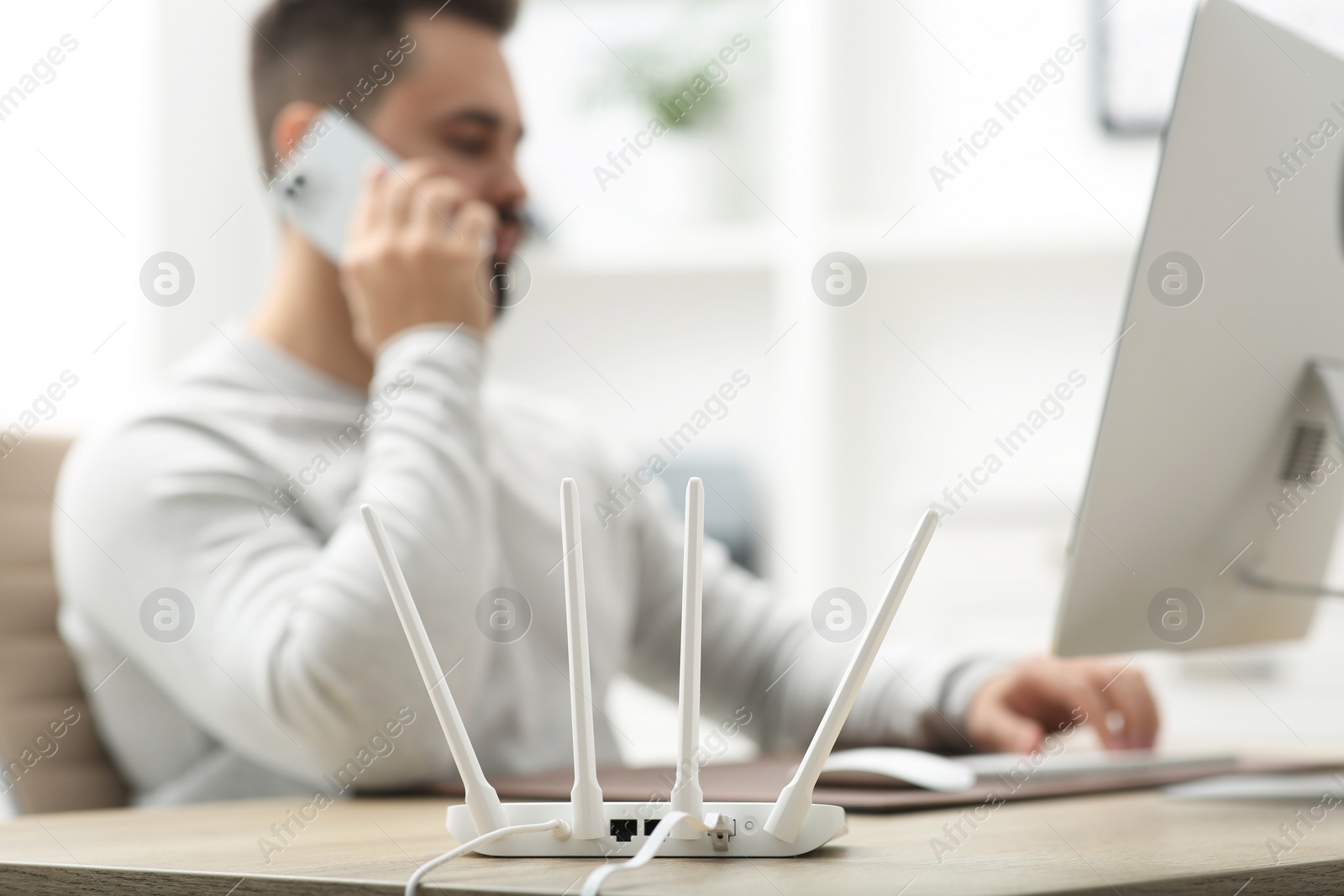 Photo of Man talking on smartphone while working at wooden table indoors, focus on Wi-Fi router