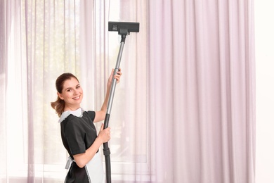 Female worker removing dust from curtains with professional vacuum cleaner indoors