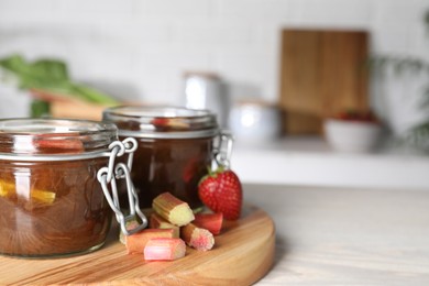 Photo of Jars of tasty rhubarb jam, cut stems and strawberry on white table. Space for text