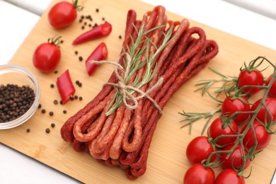 Photo of Bundle of delicious kabanosy with tomatoes and rosemary on white wooden table, closeup