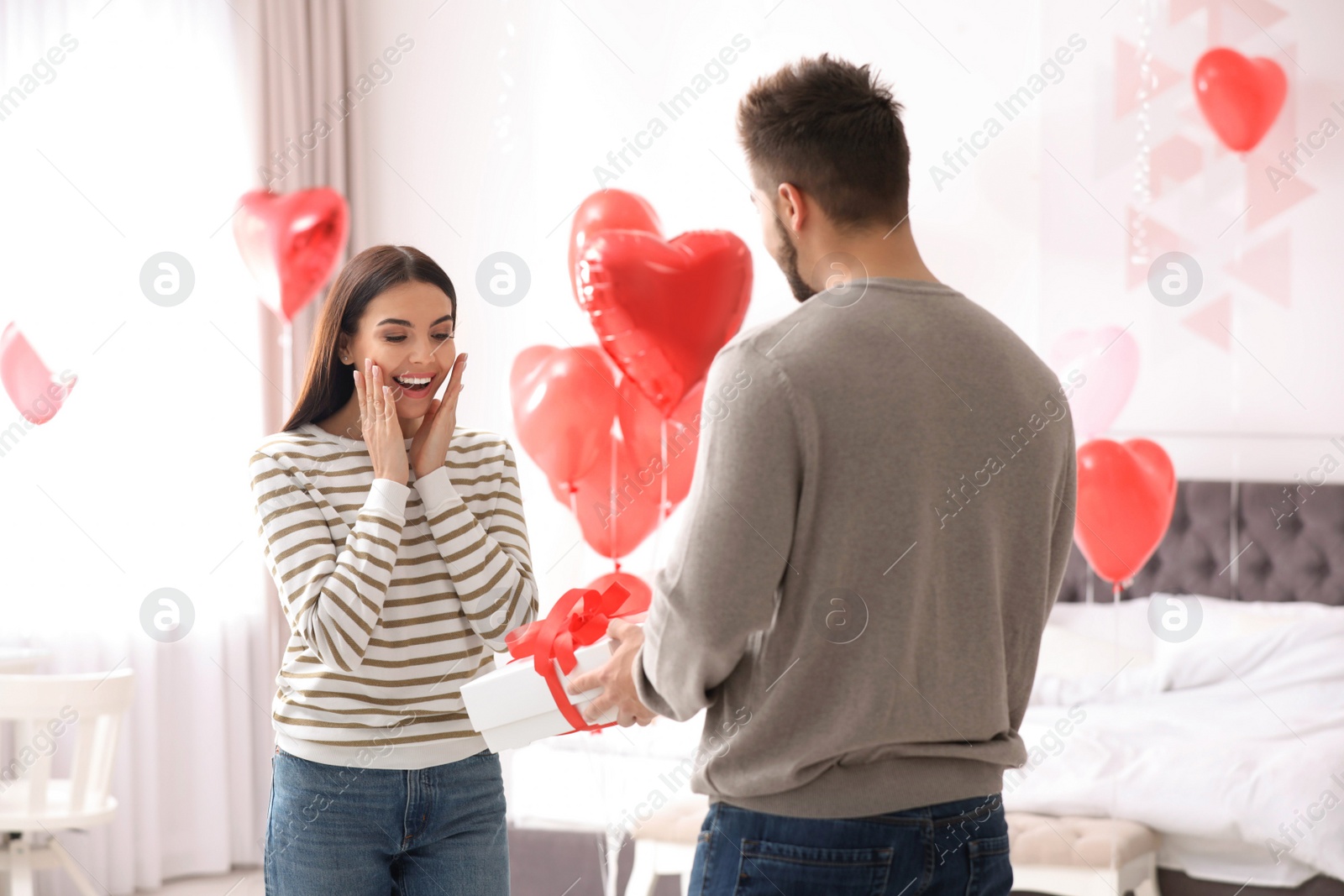 Photo of Young man presenting gift to his girlfriend in bedroom decorated with heart shaped balloons. Valentine's day celebration