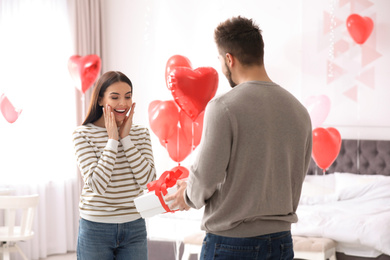 Photo of Young man presenting gift to his girlfriend in bedroom decorated with heart shaped balloons. Valentine's day celebration
