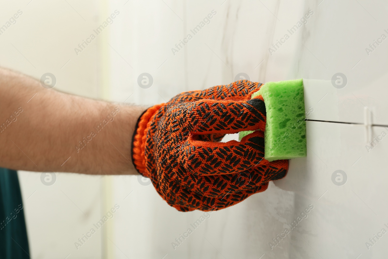 Photo of Man cleaning joint between ceramic tiles with sponge on wall, closeup. Building and renovation works