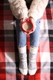 Photo of Woman relaxing with cup of hot winter drink on checkered plaid, top view