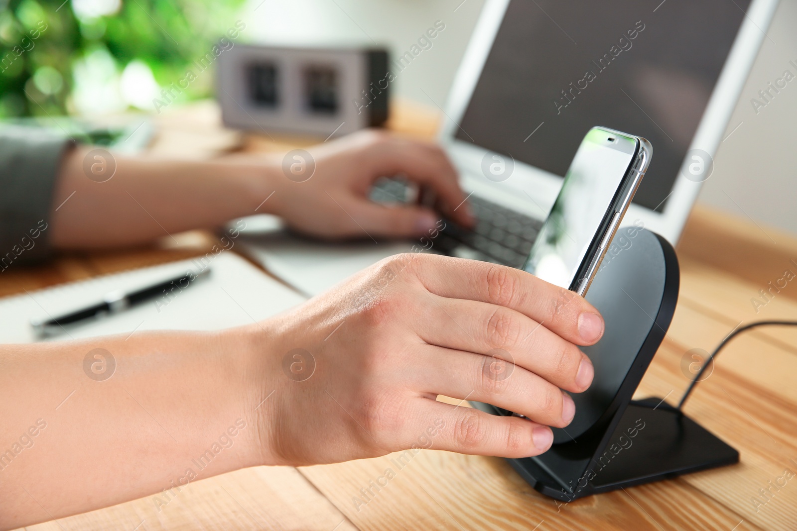 Photo of Man putting mobile phone onto wireless charger at wooden table, closeup. Modern workplace accessory