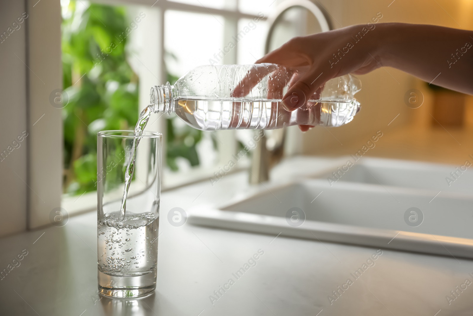 Photo of Woman pouring water from bottle into glass in kitchen, closeup