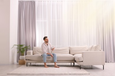 Photo of Happy man drinking coffee while resting on sofa near window with beautiful curtains in living room