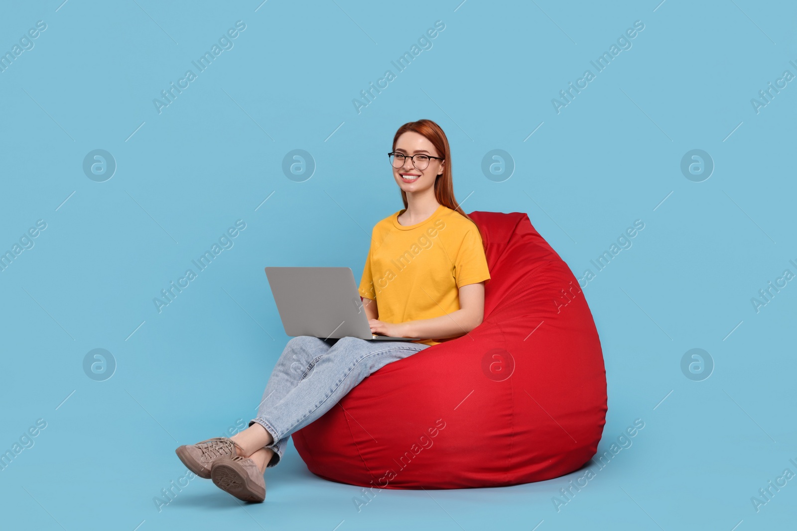 Photo of Smiling young woman with laptop sitting on beanbag chair against light blue background