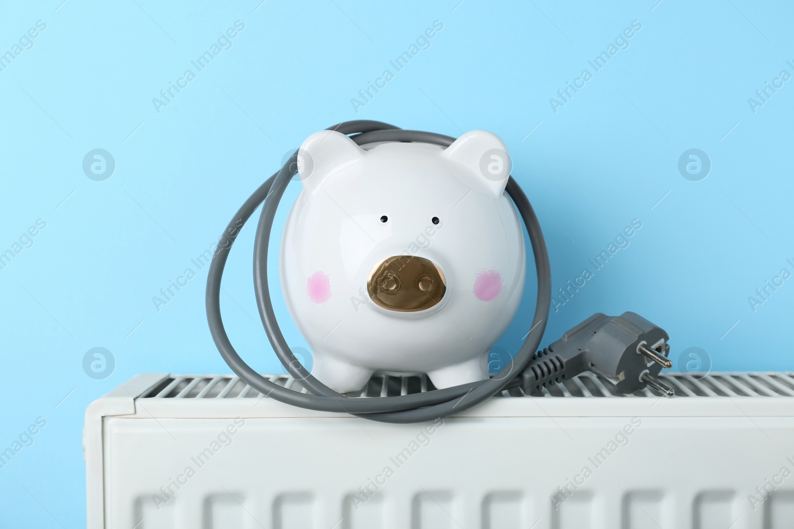 Photo of Piggy bank with plug on heating radiator against light blue background