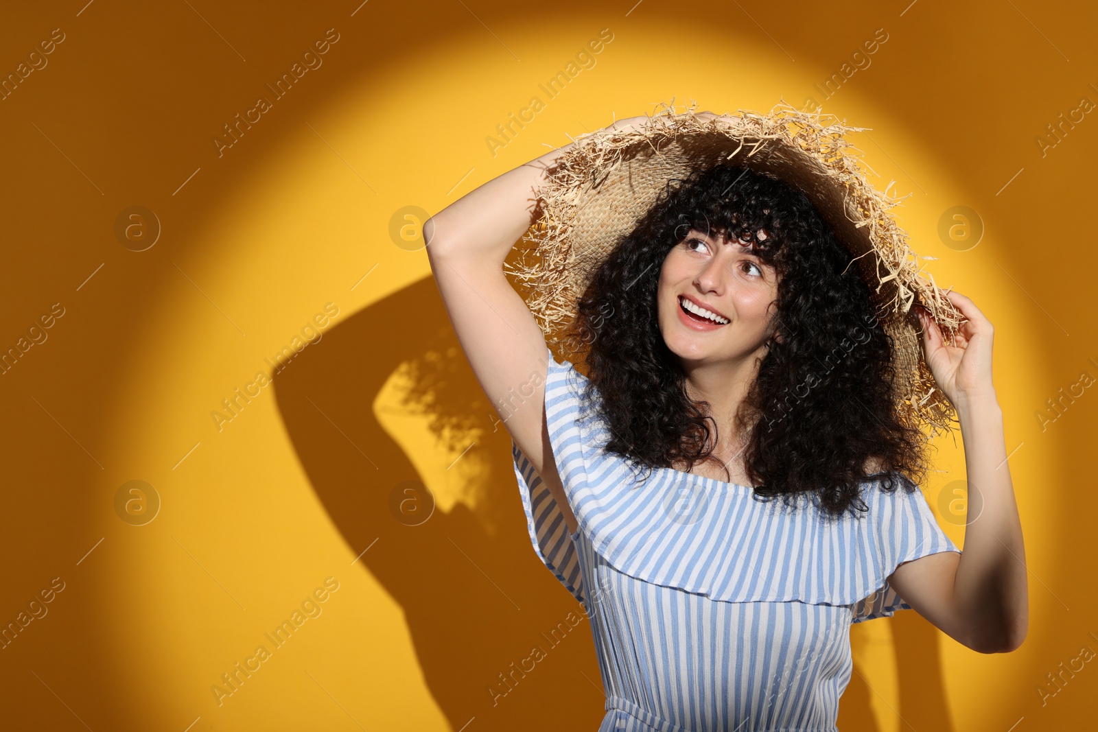 Photo of Beautiful young woman wearing straw hat in sunlight on orange background, space for text. Sun protection accessory
