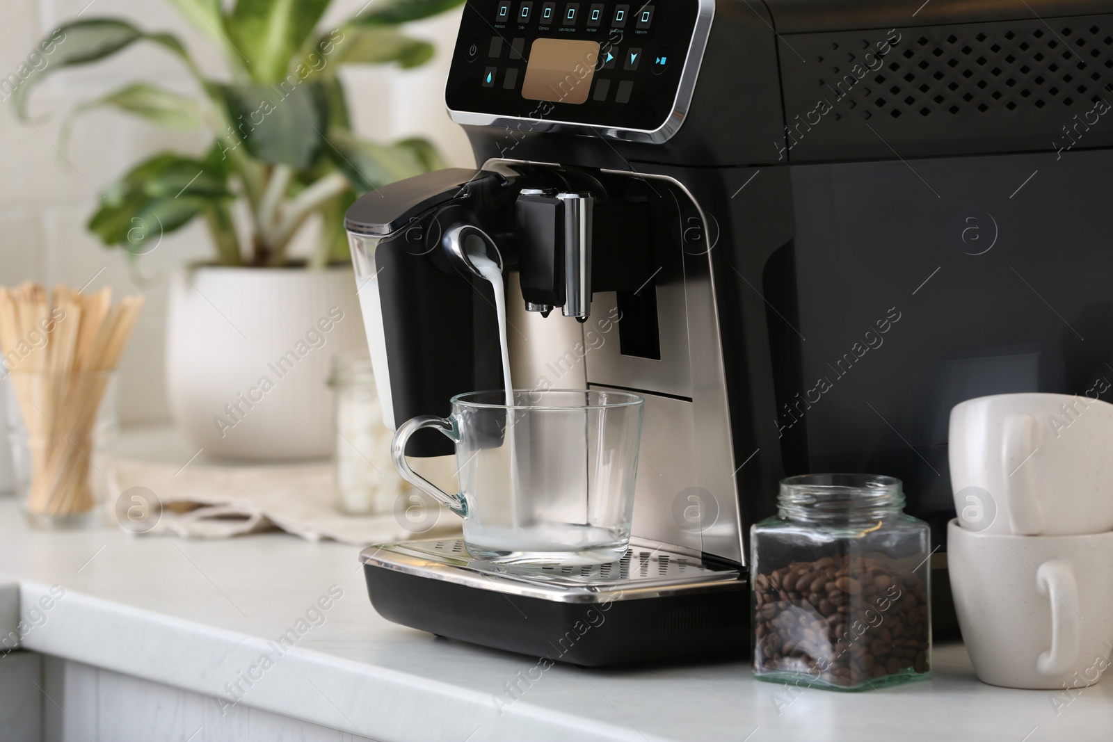 Photo of Modern coffee machine pouring milk into glass cup on white countertop in kitchen