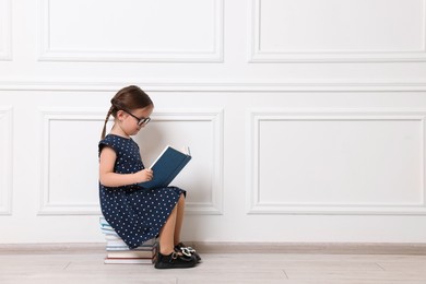 Photo of Cute little girl reading on stack of books near white wall. Space for text