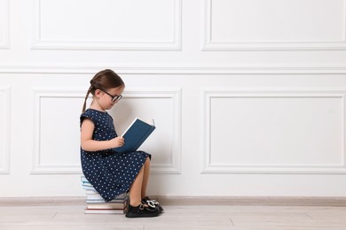 Cute little girl reading on stack of books near white wall. Space for text