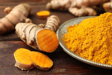 Photo of Plate with aromatic turmeric powder and cut roots on wooden table, closeup