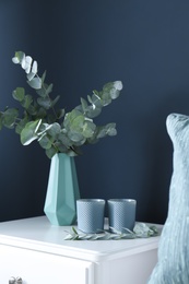 Photo of Beautiful eucalyptus branches and candles on white table near blue wall. Interior element