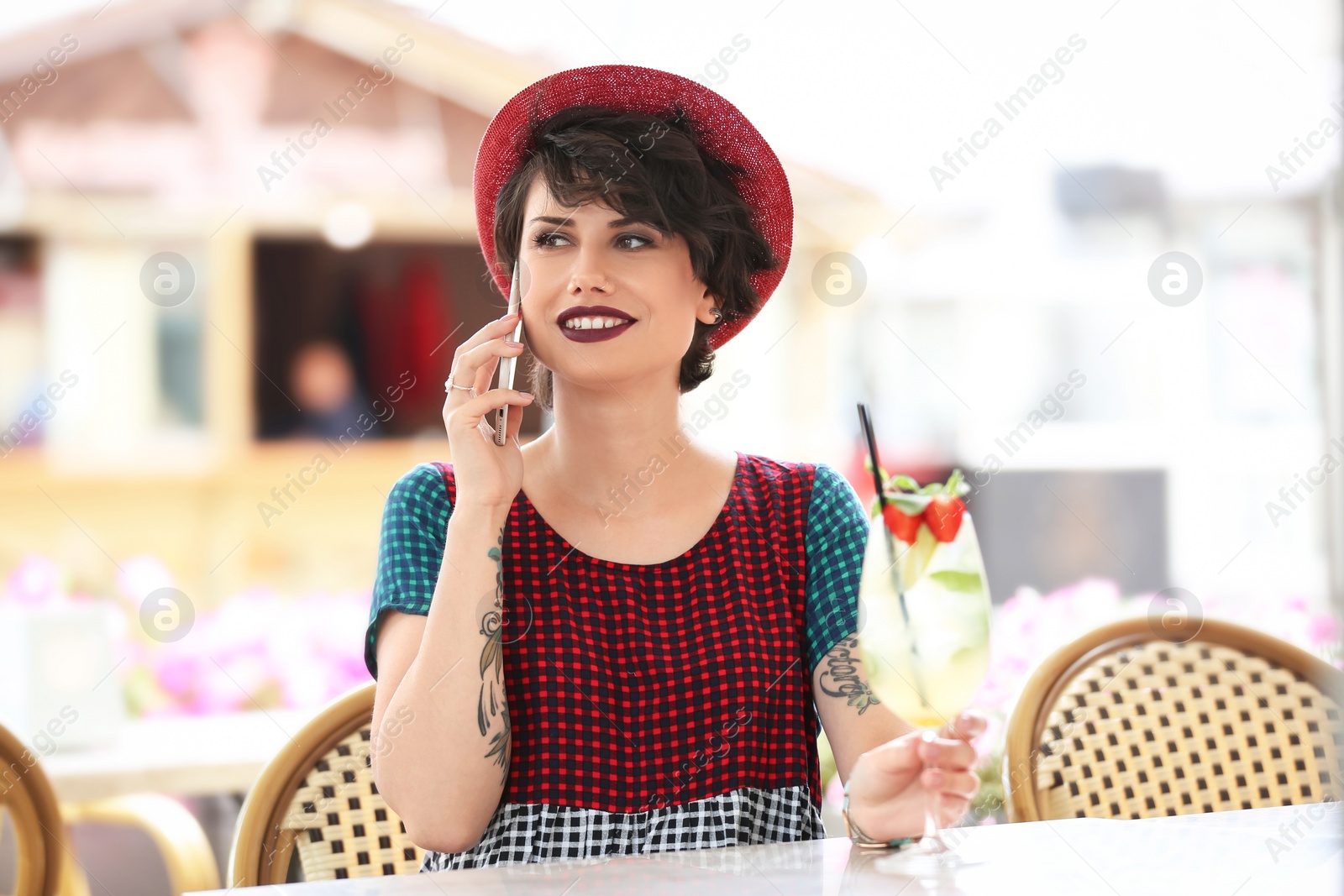 Photo of Young woman with glass of tasty lemonade talking on phone in open-air cafe