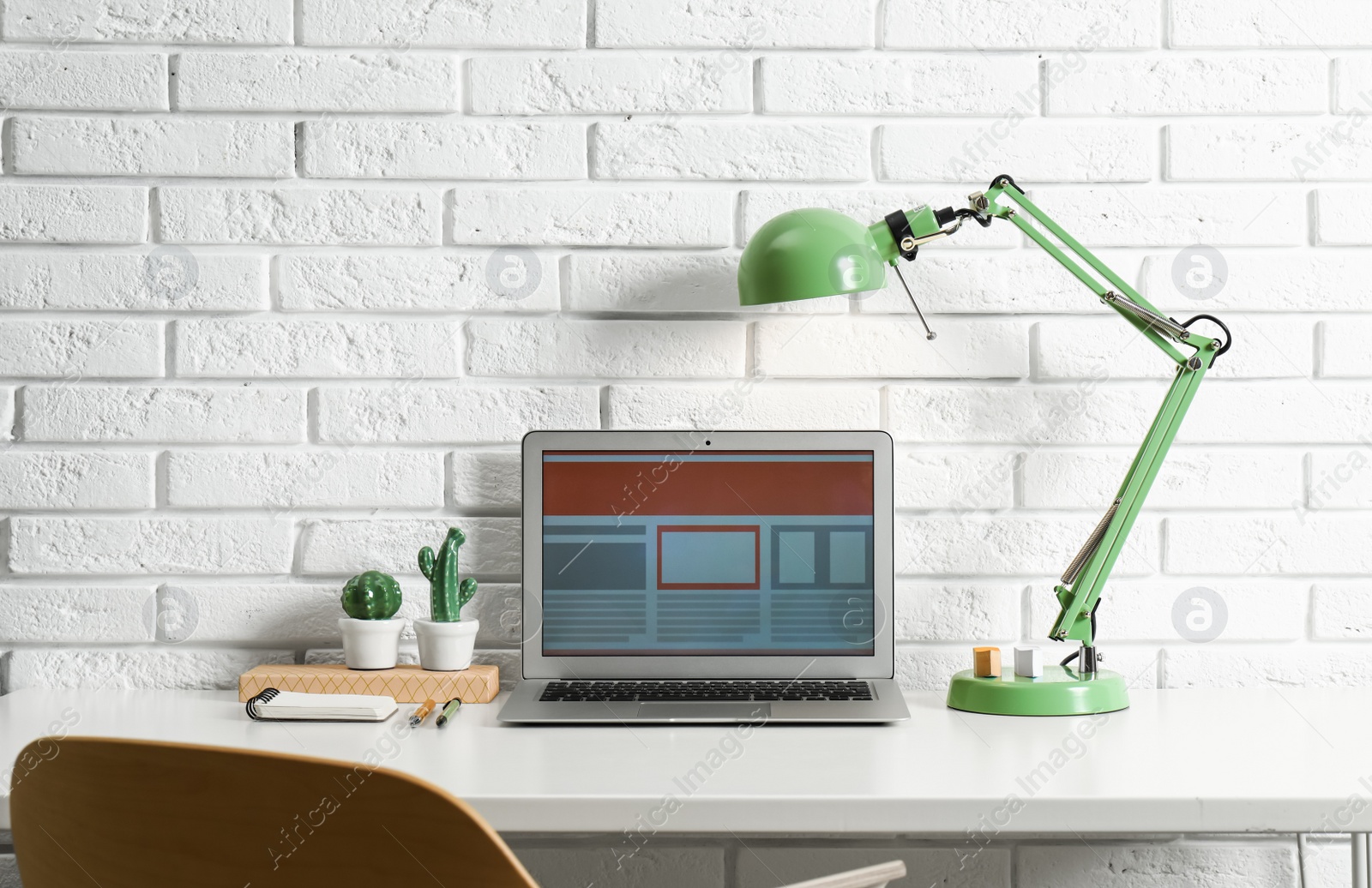 Photo of Modern workplace with laptop and lamp on table near brick wall. Space for design